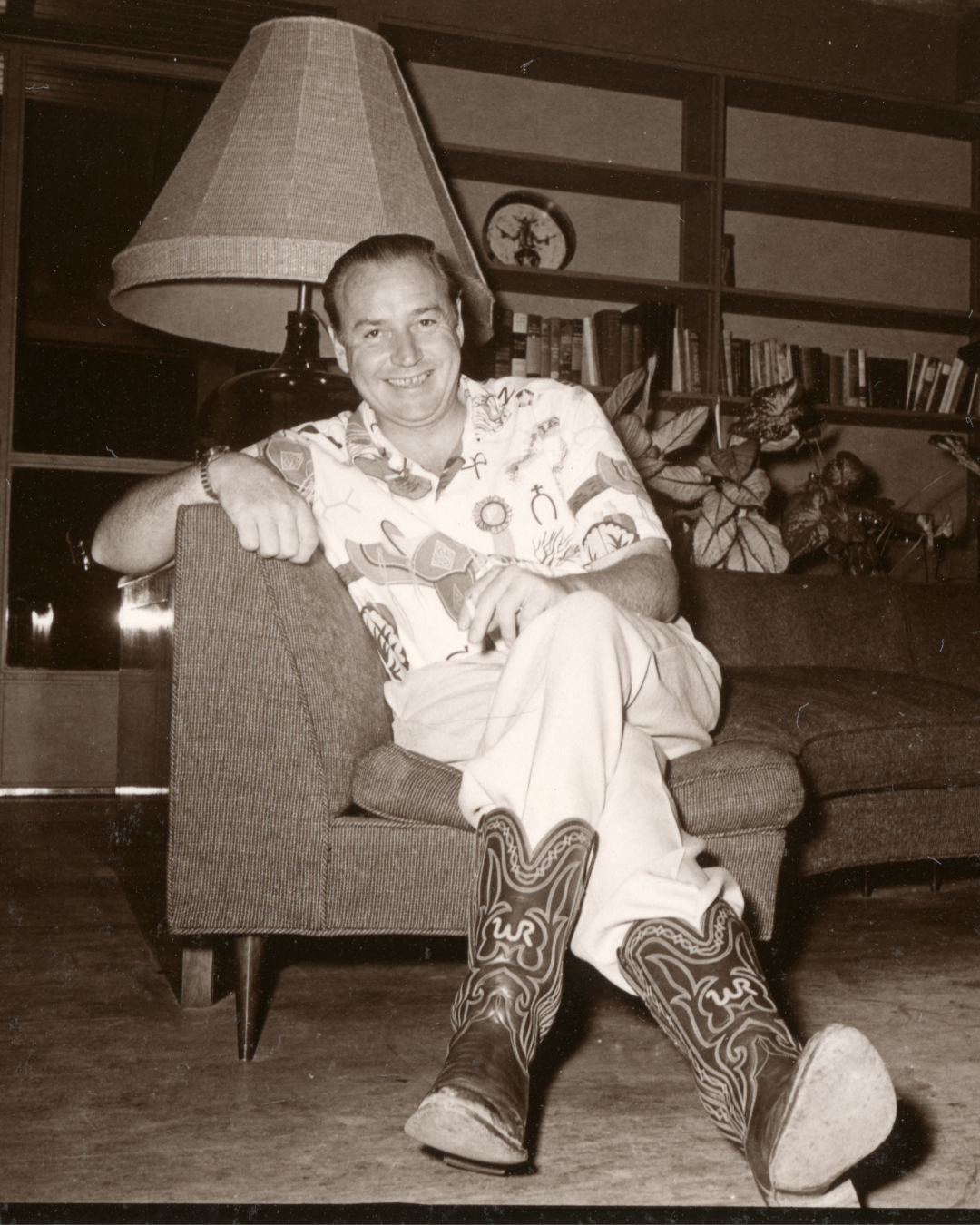 Winthrop Rockefeller sitting in on the couch in his home. He's smiling and his legs are crossed. His cowboy boots display his signature "WR" logo.